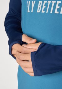 Long-sleeve athletic shirt in blue with dark blue sleeves, featuring a thumbhole design and a white "FLY BETTER" text across the chest.