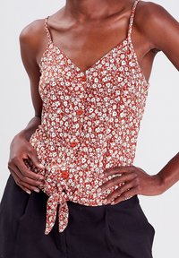 Blouse sans manches à motifs floraux, avec un col en V, fond marron, fleurs blanches et boutons rouges ; nœud noué à la taille pour plus de détails.