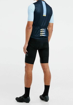 Man draagt lichtblauw en marineblauw wielertenue met zwarte fietsbroek en zwarte schoenen, van achteren gezien tegen een witte achtergrond.