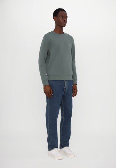 Sudadera de algodón verde con cuello redondo, puños y dobladillo acanalados. Combinada con jeans de mezclilla azules y zapatillas blancas. Diseño simple y moderno.