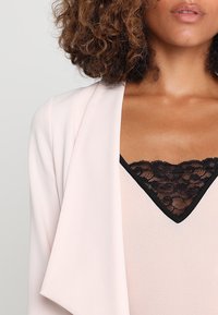 Světle růžový blazer s volným designem, který má černou krajkovou detail na výstřihu. Měkká tkanina a otevřený styl vpředu jsou patrné.
