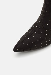 Bout pointu en daim noir orné de petits clous argentés répartis uniformément sur la surface sur un fond blanc.