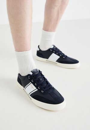 Sötétkék sportcipők fehér díszítéssel, velúr és textil felsőrész, fűzős kialakítás, enyhén textúrázott gumitalppal, a nyelvén logóval ellátva.