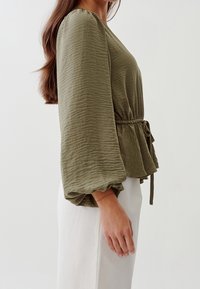 Blusa verde oliva con lunghe maniche a sbuffo, tessuto testurizzato, vita elastica con dettaglio di tie, indossata con una gonna di un leggero color crema.