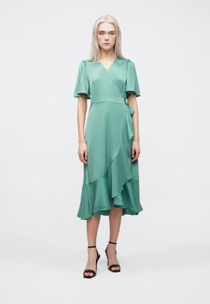 Grön omlottklänning med korta, utsvängda ärmar och en lager-på-lager, asymmetrisk fåll. Lättviktig tyg med en slät yta. Svarta remsandaler.