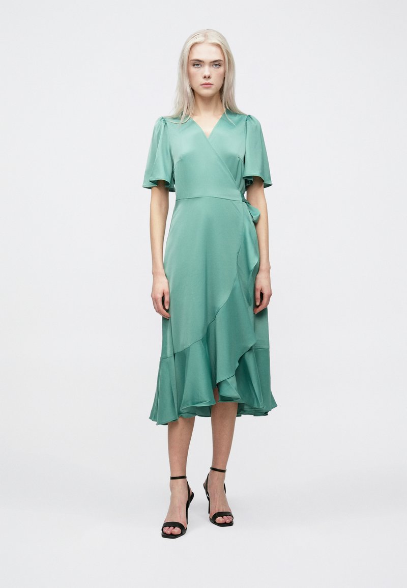 Πράσινο φόρεμα με φουσκωτά, κοντά μανίκια και στρωτή, ασύμμετρη κάτω πλευρά. Ελαφρύ ύφασμα με ομαλή υφή. Μαύρες λουρίδες τακούνια.