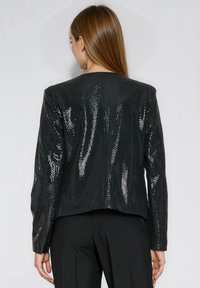 Kobieta ubrana w czarną cekinową kurtkę i czarne spodnie, pokazana od tyłu na neutralnym tle.