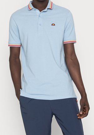 Hombre con camisa tipo polo azul claro con cuello y mangas a rayas en rojo, naranja y amarillo, combinada con pantalones azul oscuro.