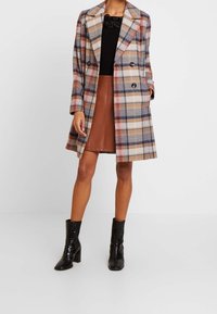 Dubbelt knäppt rutigt ytterplagg i framträdande färger som beige, marinblå och rost, med bred krage och långa ärmar. Matchas med en brun imitationläderskjol och blanka svarta ankelstövlar.