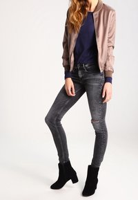 Veste bomber en satin taupe clair, pull marine, jean skinny gris foncé déchiré, bottines noires à texture lisse. Modèle en position debout.