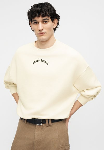 Άνδρας με σγουρά σκούρα μαλλιά, φοράει εκρού φούτερ Palm Angels και καφέ παντελόνι με μαύρη ζώνη, τα χέρια του ακουμπούν στους γοφούς.