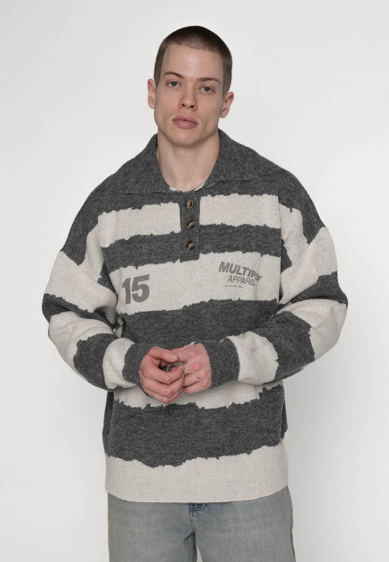 Jeune homme aux cheveux courts portant un pull rayé gris et blanc avec des boutons et les inscriptions "15" et "MULTIPLY APPAREL", debout devant un fond uni.