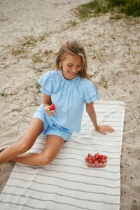 Femme en tenue bleue assise sur une couverture rayée sur le sable, tenant et souriant à une fraise avec un récipient de fraises à proximité.