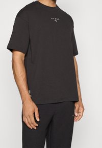 Camiseta de algodón negra con mangas cortas. Presenta texto impreso "HYROX" y un logo en el pecho. Corte recto y textura suave.