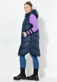 Marinblå puffväst med huva, med kviltat mönster, sidofickor och asymmetrisk nederkant, bärs över en lila långärmad topp och blå jeans.