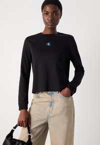 Junge Frau, die ein schwarzes langärmliges Cropped-T-Shirt mit blauem "cK"-Logo und hellen, hochgeschnittenen Jeans trägt und eine schwarze, pelzige Handtasche hält.