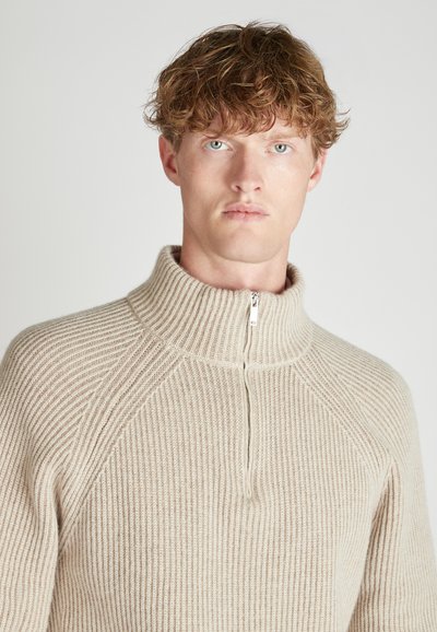 Beige gerippter Pullover mit hohem Kragen und halbem Reißverschluss, aus strukturiertem Strickstoff gefertigt, verfügt über Raglanärmel für eine figurbetonte Silhouette.