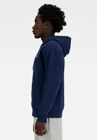 Navy hooded sweatshirt made of cotton blend, featuring a central kangaroo pocket and ribbed cuffs, worn with gray sweatpants.