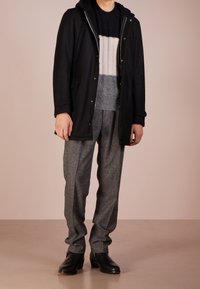 Cappotto nero con cappuccio sopra un maglione a righe grigie e crema, abbinato a pantaloni grigi strutturati e stivali in pelle nera.