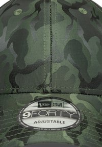 Zelená čiapka s maskáčovým vzorom vyrobená z tkaniny, s nastaviteľným popruhom a okrúhlou nálepkou označenou "NEW ERA 9FORTY ADJUSTABLE".