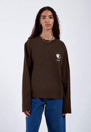 T-shirt a maniche lunghe marrone con scollatura rotonda. Presenta una grafica di un cuore bianco sul lato sinistro del petto. Taglio semplice e rilassato. Indossato con jeans blu.
