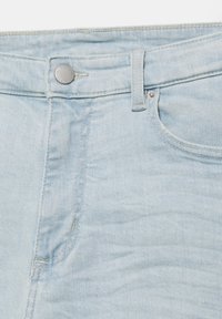 Jean en denim bleu clair montrant la ceinture avant, un bouton en métal, une passants de ceinture et une poche avant partielle avec des détails de couture.