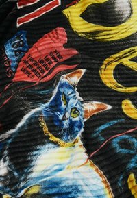 Schwarzer Stoff mit einem Aquarell-Katzendesign in Blau und Orange, mit gelben Augen und einem goldenen Halsband. Helle Farbakkente und sichtbarer Text.