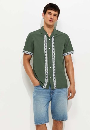 Joven con camisa verde de manga corta con ribete blanco estampado y pantalones cortos vaqueros azul claro, de pie con una mano en el bolsillo.