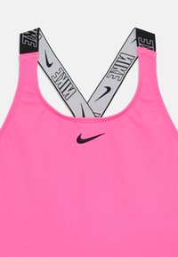 Rosa sports-BH med mykt stoff; har svart Nike-logo og grå, svartprikkede krysse-stropper. Glatt tekstur, tettsittende design.