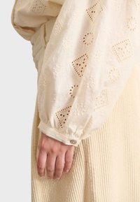 Lichtcrème katoen blouse met geborduurde bloemenpatronen, voorzien van een knoopmanchet. Overlay op een gestructureerde beige ribstof.