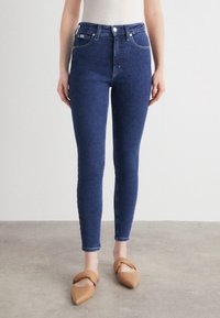 Jeans ajustados en denim azul oscuro con una cintura alta, que cuentan con un cierre de botón frontal y un diseño clásico de 5 bolsillos. Textura suave.