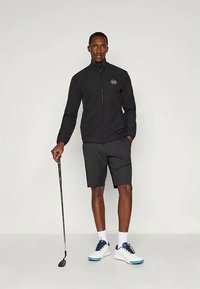 Homem com uma jaqueta preta e calções, segurando um taco de golfe, de pé com uma mão no bolso, usando ténis brancos com atacadores azuis.