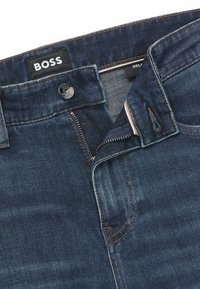 Mörkblå denimjeans med dragkedja och knappstängning, med en slät textur, kontrasterande orange sömmar och ett varumärkesetikett vid midjan.