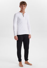 Hvid langærmet henley-skjorte med knappet halsudskæring og sorte pyjamabukser. Skjorten har en tekstureret stofkvalitet med et subtilt mønster.