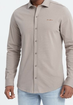 Homme portant une chemise beige à manches longues avec un petit texte brodé sur la poitrine, associée à un jean bleu clair.
