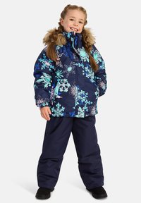 Marinblå pufferjacka med flerfärgade snöflingemönster och en huva med fuskpälsfoder, kombinerad med matchande marinblå snöbyxor och svarta stövlar.