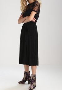 Svart plisserad midiklänning med genomskinlig spetsoverdel vid axlarna, kombinerad med blommönstrade ankelskor med klack.