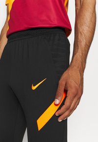 Czarne spodnie sportowe z teksturowanym pasem, z małym pomarańczowym logo Nike i wyraźnym pomarańczowym akcentem po bokach.