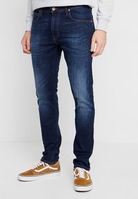 Homme portant un jean slim bleu foncé et des baskets en daim couleur moutarde avec des lacets blancs, debout devant un fond blanc uni.