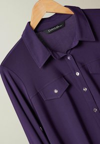 Purple long-sleeve shirt with a classic collar, two chest pockets, and silver buttons. Made from smooth, textured fabric.