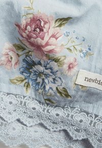 Vaaleansininen kangas, jossa on vaaleanpunaisia ja sinisiä kukkakuoseja, sekä herkällä pitsireunalla alareunassa ja valkoisella lapulla, jossa lukee "newbie".