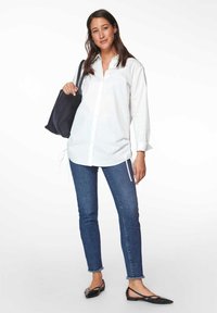 Weißes Hemd mit gerafften Seiten, blaue enge Jeans mit ausgefransten Säumen und schwarze flache Schuhe, kombiniert mit einer marineblauen Tragetasche.