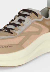 Μπεζ και καφέ αθλητικό παπούτσι Calvin Klein με σκούρα καφέ κορδόνια, επάνω μέρος από σουέντ και δίχτυ, χοντρή λευκή σόλα και λογότυπο στην πλευρική επιφάνεια.