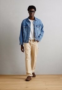 Denim jakna, bela majica in bež hlače; model ima kodrasto lase in očala; rjave natikače; nevtralen ozadje.