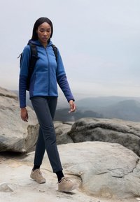 Blaue und marinefarbene Zip-Jacke, graue Hose und beige Wanderschuhe. Geht über felsiges Gelände mit einem Rucksack. Weicher Stoff, sportive Passform.