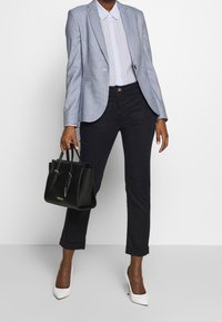 Blazer a quadri blu chiaro sopra una camicia blu chiaro, pantaloni neri, tacchi bianchi e una borsa nera con accessori in oro.