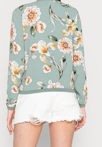 Suéter floral verde con flores naranjas y blancas, con mangas largas y dobladillo acanalado, combinado con pantalones cortos blancos desgastados.