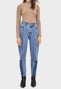 Kvinna iklädd högmidjade, tvåfärgade blå jeans, en figurnära beige ribbstickad tröja och svarta spetsiga ankelkängor mot en vit bakgrund.