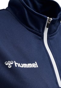 Giacca zip-up blu navy con texture a coste; presenta un collo alto e una zip bianca, con un grande logo "hummel" bianco sul lato sinistro.