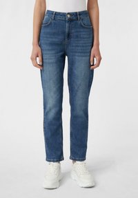Blaue hochgeschnittene Jeans mit geradem Bein, fünf Taschen und einem vorderen Knopfverschluss. Getragen mit weißen Sneakers.